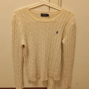 Vit kabelstickad tröja Polo Ralph Lauren - Kabelstickad vit tröja från Polo Ralph Lauren med klassisk blå logga broderad på bröstet. Tröjan har rund halsringning, ribbade muddar och är långärmad. Perfekt för dig som gillar en clean och tidlös stil. Jag köpte den i deras affär i Stockholm 