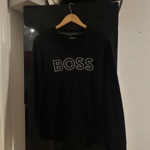 Svart långärmad tröja från BOSS - Svart långärmad tröja från BOSS med vit broderad logga framtill. Tröjan är i storlek M men passar även S, köptes på NK för något år sedan men har inte används så mycket. Säljs pga den ej passar min stil längre. 
