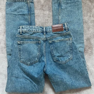 Blåa Jeans Från Zara - Zara jeans i mellanblå tvätt med raka ben och hög/mellan midja. Jeansen har fickor, gylf och läderpatch bak i midjan. Perfekta för en avslappnad och tidlös look.