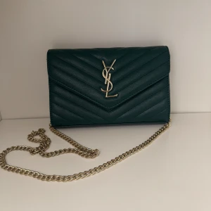 Grön quiltad YSL kuvertväska i skinn - Säljer en snygg grön kuvertväska från YSL i quiltat skinn med guldigt kedjeaxelband och ikonisk YSL-logga framtill. Väskan har ett vinrött innerfoder och flera fack, perfekt för det viktigaste. Stilren och lyxig accessoar som lyfter varje outfit. Inte äkta