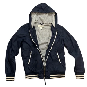 Mörkblå vindjacka med huva - Snygg mörkblå vindjacka med grått foder och huva. Jackan har dragkedja och ribbade muddar i vitt och blått. Perfekt för blåsiga dagar. Har ett litet hål men de syns inte så mycket den är perfekt till våren eller kalla sommar dagar 