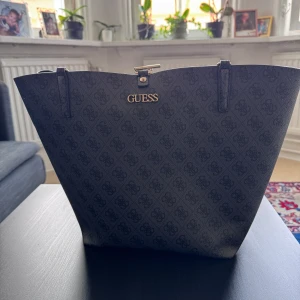 Guess shoulderbag - Snygg svart shoulderbag från Guess med diskret mönster och guldfärgade detaljer. Väskan har två långa axelband och stängs med en cool guldfärgad kedja lås. Perfekt storlek för att få plats med allt du behöver. Klassisk och stilren design. Nypris ca 1300kr mitt pris 999kr