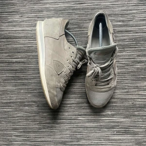 Grå sneakers från Phillipe model Trainers - Snygga grå sneakers från Philippe Model Trainers i storlek 40. Skorna har en stilren design med rund tå, snörning och platt sula. Ovandelen är i mocka och textil, vilket ger en soft vibe och skön känsla. Perfekt för dig som gillar minimalistisk stil och neutrala färger. Passar 42 säkert.