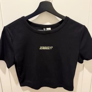 Svart croppad topp H&M Seriously? - Svart croppad topp från H&M med trycket 'SERIOUSLY?' i grönt framtill. T-shirten har rund halsringning och korta ärmar. Perfekt för dig som gillar en chill och edgy stil.