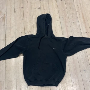 Svart Nike hoodie med huva - Svart hoodie från Nike med klassisk logga på bröstet. Tröjan har huva med snörning och en stor magficka framtill. Perfekt för chill dagar eller när du vill ha en enkel och stilren look. Materialet är mjukt och känns skönt mot huden.