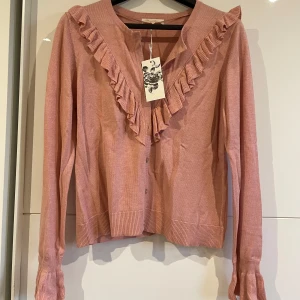 Rosa kofta med volang från RoseBud - Supersöt rosa kofta från RoseBud med volangdetaljer längs halsringningen och knappar framtill. Helt ny.