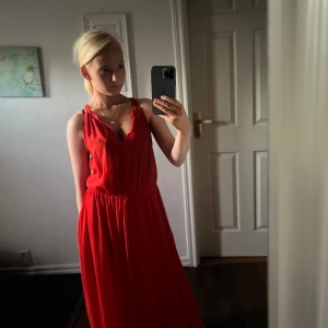 Röd långklänning med v-ringning - Säljer en snygg röd långklänning med v-ringad hals och smala axelband. Klänningen har ett lätt och luftigt tyg som faller fint och ett resår i midjan för skön passform. Perfekt för dig som gillar färgstarka och stilrena plagg.