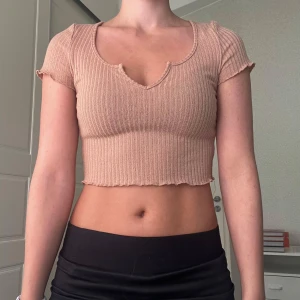Beige ribbad croppad topp - Säljer en beige croppad topp med ribbad struktur och korta ärmar. Toppen har en snygg v-ringning med liten cutout och vågig kant nertill och vid ärmslut. 