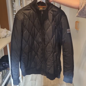 Svart quiltad bomberjacka från BOSS - Svart bomberjacka från BOSS med quiltad framsida och ribbade muddar. Jackan har dragkedja, lång ärm och snyggt BOSS-märke på ärmen. Materialet är polyester och jackan har en stilren, normal passform. Perfekt för dig som vill ha en clean look med lite edge.