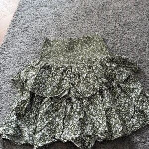 Grön blommig volangkjol Designers Remix - Supersöt grön kortkjol från Designers Remix med vitt blommigt mönster. Kjolen har smockad midja och flera lager med volanger som ger en flowy och chill vibe. Perfekt för dig som gillar att sticka ut med detaljer och mönster.