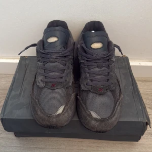 New Balance 2002R mörkgrå sneakers - Säljer ett par New Balance 2002R sneakers i mörkgrått med slitna detaljer och mesh-paneler. Skorna har grov mocka, rund tå och platt sula. Insidan har gul toning och klassisk N-logga på sidan. Perfekt för dig som gillar streetwear och chunky sneakers.