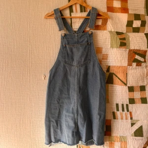 Blå jeansklänning med hängslen - Säljer en chill blå jeansklänning med breda hängslen och stor ficka framtill. Klänningen har en loose passform och rå kant nedtill för en avslappnad vibe. Perfekt att styla med t-shirt eller hoodie under. Klassisk denimlook som funkar året runt.