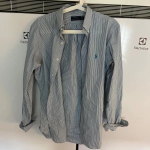 Randig skjorta från Polo Ralph Lauren - Klassisk skjorta från Polo Ralph Lauren i blåvitt randigt mönster. Skjortan har lång ärm, knappar framtill och den ikoniska broderade loggan i blått på bröstet. Tillverkad i mjuk bomull och har en normal passform. Perfekt för en clean och stilren look.