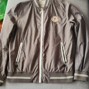 Brun bomberjacka från Moncler - Snygg brun bomberjacka från Moncler med klassisk logga på bröstet. Jackan har vita detaljer vid dragkedjor och muddar, samt ribbade kanter. Tillverkad i lätt polyester, perfekt för vår och höst. Stilsäker och enkel att matcha med olika outfits. Storlek S kanske M 