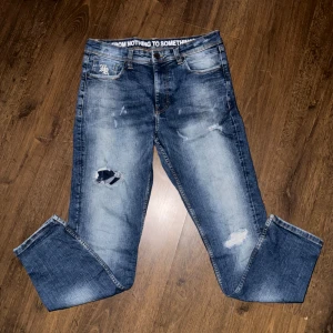 Blå slitna jeans från Hoodrich, 32W - Snygga blå jeans från Hoodrich med slitningar och hål på benen för en streetstyle-look. Jeansen har broderad logga på fickan, klassisk femficksdesign och normal passform. Materialet är jeans med en tvättad finish och coola detaljer i midjan.