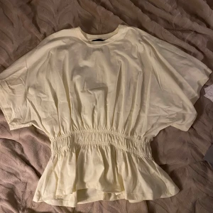 Ljusgul blus med resår i midjan - Superfin ljusgul blus med bred resår i midjan som ger en snygg volangkant nedtill. Blusen har korta, vida ärmar och rund halsringning. Perfekt för dig som gillar en avslappnad men ändå söt stil.