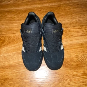 Adidas Samba OG svarta sneakers mocka - Svarta Adidas Samba OG sneakers i mocka med klassiska mörk gröna ränder på sidorna. Tjock svart sula, svart snörning och guldfärgad Samba-text på sidan. Perfekt för dig som gillar retrostil och streetwear.