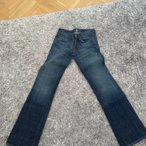 Bootcut jeans från 7 For All Mankind - Mörkblå bootcut jeans från 7 For All Mankind med snygg tvätt och klassiska kontrastsömmar. Bakfickorna har en glittrig detalj i form av stenar. Jeansen är i klassisk denim och har normal passform med medelhög midja.