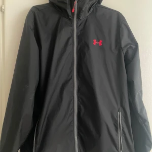 Svart vindjacka från Under Armour - Svart vindjacka från Under Armour med huva och hel dragkedja framtill. Jackan har två fickor med dragkedja och en röd logga på bröstet. Tillverkad i lätt syntetmaterial, perfekt för blåsiga dagar. Enkel och sportig design som funkar till det mesta.