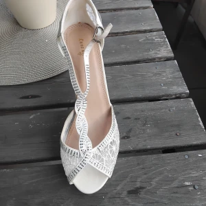 Vita spetsiga pumps med strassdetaljer - Eleganta vita pumps med hög klack, dekorerade med glittrande strass och vacker spets. Skorna har öppen tå och smala remmar som korsas framtill. Perfekta för dig som vill sticka ut med en lyxig och feminin look.