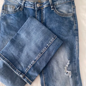 Blå jeans med slitning på benet - Snygga blå jeans med klassisk femficksdesign och en cool slitning på ena benet. Jeansen har normal passform och är tillverkade i mjukt denimtyg med ljusa tvättade partier. Perfekta för en avslappnad och trendig look.