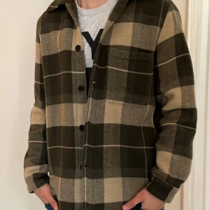 Rutig overshirt från A Day's March - Snygg rutig overshirt från A Day's March i mörkgrön och beige. Jackan har klassisk krage, bröstficka och knäppning med svarta knappar. Materialet är en mix av ull och polyamid med bomullsfoder, perfekt för lager-på-lager. Passar dig som gillar stilren och chill look.