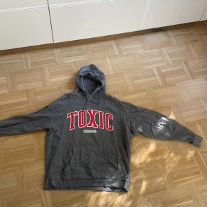 Grå hoodie  XL - Grå hoodie från Vailent i storlek XL med stor röd TOXIC-text på bröstet och vit detalj under. Klassisk känguruficka, huva med snörning och långa ärmar. Perfekt till hösten 