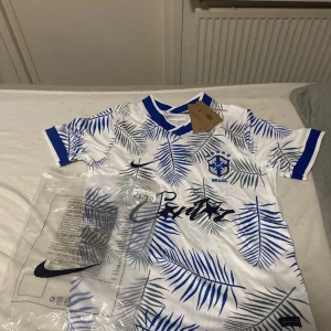 Vit och blå Brasilien fotbollströja Nike - Snygg Brasilien fotbollströja från Nike i vitt med blå palmbladsmönster och blå detaljer vid krage och ärmslut. Tröjan har korta ärmar och Brasil-logga på bröstet. Perfekt för dig som vill sticka ut på planen eller läktaren.