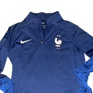 Blå långärmad Nike Frankrike Dri-Fit - Snygg marinblå långärmad fotbollströja från Nike med Frankrikes landslagsemblem och två stjärnor på bröstet. Tröjan har halv dragkedja, slim fit och är gjord i Dri-Fit material som andas. Ärmarna har diskret mönster i blått för extra stil.