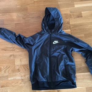 Svart vindjacka från Nike med huva - Snygg svart vindjacka från Nike med huva och dragkedja framtill. Jackan har två praktiska fickor med dragkedja och en vit Nike-logga på bröstet. Perfekt för blåsiga dagar och enkel att matcha med streetwear. Lätt och smidig modell.