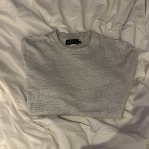 Grå stickad tröja från Riley - En stilren grå stickad tröja från Riley med rund hals och långa ärmar. Tröjan har en enkel design utan mönster, ribbade muddar vid ärmslut och nederkant. Perfekt för dig som gillar en clean och chill look.