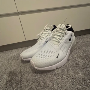 Nike Air 270 vita sneakers - Nike Air 270 sneakers i fräsch vit färg med svarta detaljer och synlig Air-sula. Ovandelen är i ventilerande syntetmaterial med snörning och rund tå. Snygg design med logga på sidan och bak, perfekt för dig som gillar sportig stil och maxad komfort.
