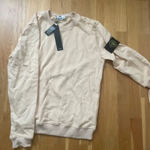 Beige sweatshirt från Stone Island - Snygg beige sweatshirt från Stone Island med klassisk rund hals och ribbade muddar. Tröjan har den ikoniska loggan på vänster ärm och är tillverkad i mjuk bomull. Perfekt för dig som gillar stilrena och trendiga plagg. Helt ny och oanvänd