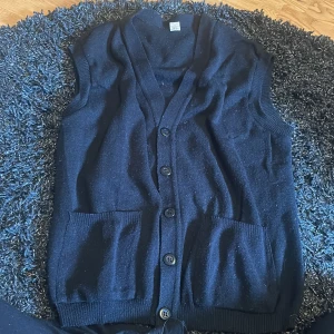 Svart stickad väst med knappar - Mysig svart stickad väst med v-ringning och knäppning framtill. Västen har två praktiska fickor och ribbade kanter vid ärmhål och nederkant. Perfekt att slänga över en t-shirt eller skjorta för en chill look.