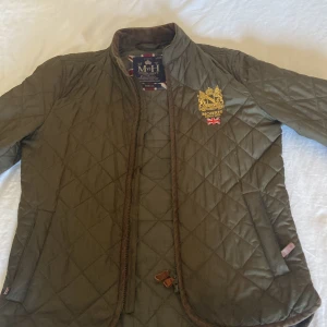 Olivgrön quiltad jacka från Morris XS - Quiltad olivgrön jacka från Morris med snyggt broderat emblem och brittisk flagga på bröstet. Jackan har dragkedja, bruna detaljer i manchester och klassisk krage. Perfekt för dig som gillar stilren och sportig look.