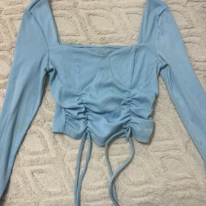 Ljusblå croppad ribbad topp - Trendig ljusblå långärmad topp med ribbad struktur och croppad passform. Toppen har snygg snörning framtill som ger en cool rynkad effekt och fyrkantig urringning. Perfekt för dig som gillar stilrena men ändå unika detaljer.