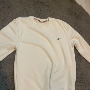 Beige sweatshirt  - Snygg beige sweatshirt  med klassisk rund hals och den ikoniska gröna krokodilloggan broderad på bröstet. Tröjan har långa ärmar och ribbade muddar. Perfekt för en clean och stilren look.
