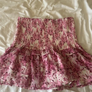 Blommig volangkjol från Gina Tricot - Supersöt kort kjol från Gina Tricot med rosa och vita blommor. Kjolen har smockad midja och flera lager volanger som ger en flowy känsla. Perfekt för dig som gillar romantisk och feminin stil.