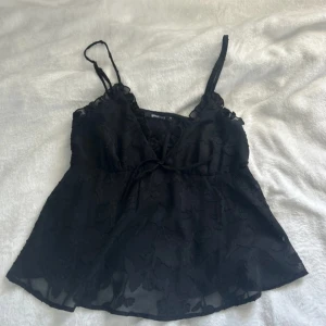 Svart blommig linnetopp - Supersöt svart linnetopp från Gina Tricot med blommigt mönster i tyget. Toppen har tunna axelband, v-ringning och en liten knytning framtill. Tunt, lätt material som känns luftigt och snyggt.