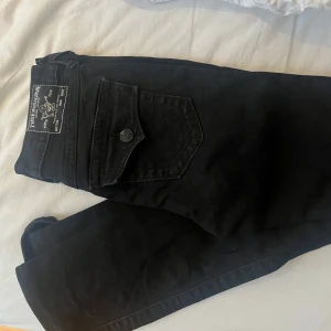 Svarta True Religion bootcut jeans - Svarta jeans från True Religion, modell Becca Mid Rise Bootcut. Snygga bakfickor med lock och knappdetalj, klassisk True Religion-lapp bak i midjan. Jeansen har bootcut-passform och är tillverkade i stretchig bomull för skön känsla.