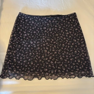 Svart blommig meshkjol med volangkant - Säljer en svart meshkjol med små blommor i rosa och lila. Kjolen är kort och har en söt volangkant längst ner som ger en extra touch. Perfekt för dig som gillar miniskirts, romantisk och trendig stil. Storlek xxs men skulle säga att den passar en Xs-S
