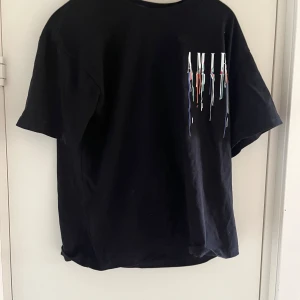 Svart Amiri t-shirt med färgstänk - Svart t-shirt från A med coolt tryck framtill där bokstäverna har färgstänk som rinner ner i blått, orange, grönt och vitt. Skön  passform och tillverkad i 100% bomull. Perfekt för dig som gillar streetwear och vill sticka ut. Använd 1-2 gånger 