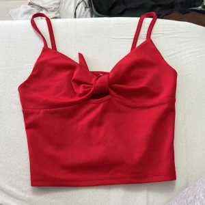 Röd croppad topp med rosett - Snygg röd croppad topp med smala axelband och en stor rosett framtill. Perfekt för dig som vill sticka ut med en färgstark och trendig look. Toppens material känns mjukt och följsamt mot huden.