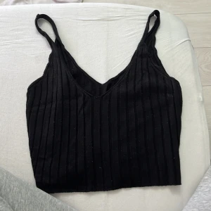 Svart ribbad linnetopp - Snygg svart croppad linnetopp med smala axelband och v-ringning. Toppen är ribbad och har en tight passform som framhäver siluetten. Perfekt att matcha med jeans eller kjol för en trendig look.