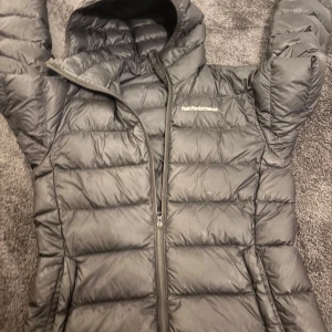 Svart dunjacka Peak Performance S - Säljer en svart dunjacka från Peak Performance i storlek S. Jackan har huva, quiltad design och dragkedja framtill. Perfekt för kalla dagar och har en clean look som funkar till de flesta stilar. Loggan syns på bröstet och materialet är syntet med dunfyllning. De finns tecken på användning, men inga defekter!