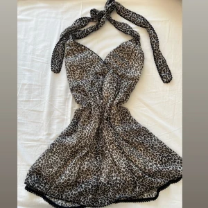 Leopardmönstrad kortklänning med knyt - Säljer en kort klänning med leopardmönster och knyt i nacken. Klänningen har omlottliv, svarta dekorativa kanter och är tillverkad i ett tunt, luftigt material. Perfekt för dig som gillar att sticka ut med djurmönster och vill ha en cool look. 🥰