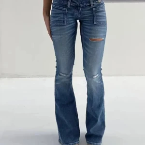 Blå bootcut jeans med slitningar - Snygga blå bootcut jeans med slitna detaljer på låren och knäna. Jeansen har låg midja och stora framfickor som ger en cool vibe. Materialet är klassisk jeans och passformen är loose med utsvängda ben. Perfekta för dig som gillar retrostil och vill sticka ut.