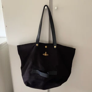 Vivienne Westwood väska - Vivienne Westwood Utility Unisex shoppingväska. Köpt för snart 3 år sen. Använde ganska mycket under ett halv år men nu har den legad orörd i 2 år. Har några sminkfläckar och mild missfärgning på insidan, vet inte om dem går bort, har själv bara testat med vatten och diskmedel. Utsidan ser så gott som oanvänd ut. Vågar ej köra den i tvättmaskinen men har damsugit, tvättat och tagit bort damm och hår så gott det går. Original pris: 3695kr 