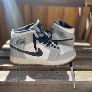 Nike Air Jordan 1 Mid grå/vit - Snygga Nike Air Jordan 1 Mid sneakers i grått och vitt med svarta detaljer och klassisk svart swoosh. Skorna har svart snörning, perforerad tå och ikoniska Air Jordan-loggan på sidan. Perfekta för dig som vill ha en clean och sportig look.