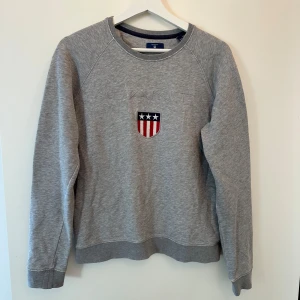 Grå sweatshirt från GANT med brodyr - Klassisk grå sweatshirt från GANT med broderad logga och sköld på bröstet.